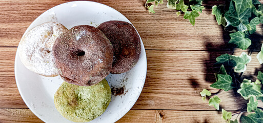 Two milk chocolate donuts, one matcha donut, and one sprinkled sugar donut are served on a white plate at a bakery. Brown background.