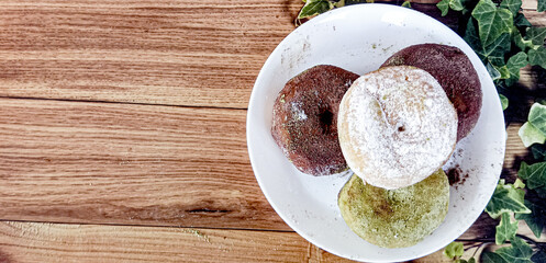 Two milk chocolate donuts, one matcha donut, and one sprinkled sugar donut are served on a white plate at a bakery. Brown background.