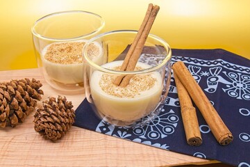 Two cups of eggnog with a cinnamon stick in one glass and cinnamon in both glasses. Pine cones and cinnamon sticks sit on a wood cutting board and blue cloth with holiday decorations. 