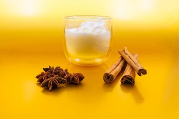 Glass of eggnog with whipped cream on top. There are star anise on one side, cinnamon sticks on the other side.  The background is metallic gold.