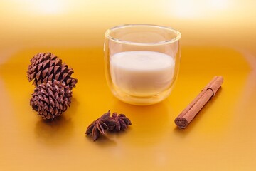 Glass of eggnog . There are pine cones on one side, cinnamon sticks on the other side and star anise in front.  The background is metallic gold.