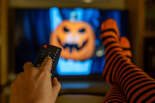 Festive Halloween streaming concept, cozy living room with striped tights, remote control in hand and unfocused TV screen in autumn atmosphere.