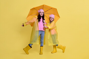 Two little cute girls in raincoats and an umbrella on a yellow background
