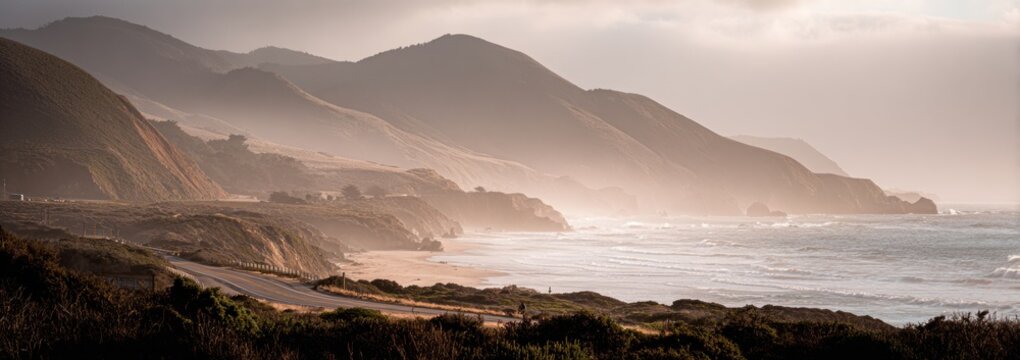 A scenic coastal highway view with rolling hills shrouded in soft, diffused sunlight, meeting the ocean with gentle waves