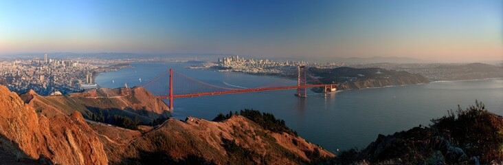 Sweeping vista of a coastal city with a large suspension bridge, taken from a high vantage point on a golden hill under a blue sky