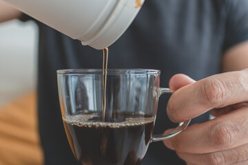 Stream of black coffee flowing from kettle to glass on wood table in soft indoor morning