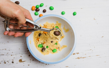 Halloween cookies, sugar cookie dough with chocolate covered peanuts candies in orange, black and green, male hand scooping raw Halloween cookie dough balls with scoop on baking sheet white background