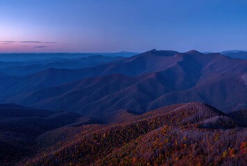 Fototapeta premium A scenic vista reveals rolling mountains layered under a deep blue and pink twilight sky, accented with foliage displaying autumn colors