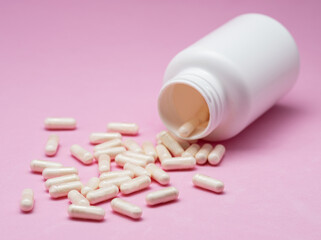 White capsules or tablets poured out of plastic bottle on pink background.