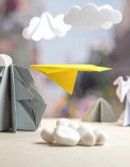 A yellow paper airplane soars above a miniature origami landscape of mountains, clouds, and other shapes on a beige surface
