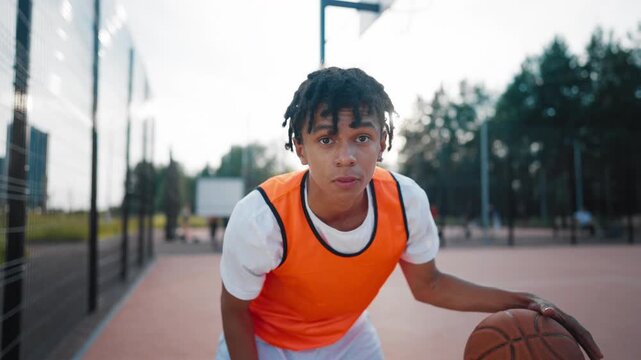 Sport and leisure for teens, black boy playing streetball outdoor, portrait. Active training with ball in open court, young african american guy looking at camera and striking ball on floor, champion