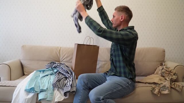 A handsome young man in a green shirt and blue jeans is sitting on the couch packing a donation box with old clothes. A young man packs clothes in a paper bag.A man admires new clothes after shopping.