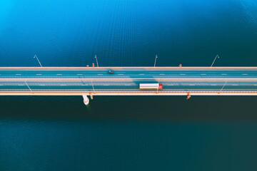 Aerial drone view of bridge asphalt road with cars and cargo trucks driving over blue river or lake water. Logistic and delivery concept.