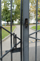 Close-up of a metal gate with intercom button restricting access to a fenced sports area in a typical city courtyard, symbolizing urban boundaries and security.