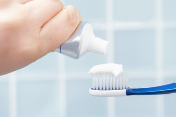 Female hand squeezing toothpaste onto toothbrush against blue tile background.