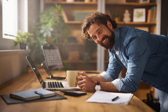 A joyful man leans on his desk in a home office, holding a cup of coffee and working intently on his laptop. Natural light fills the space, enhancing the welcoming atmosphere.