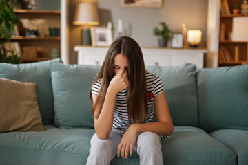 Obraz premium A teenage girl sits on a soft couch in a warmly lit living room, her head down and hand covering her face, visibly expressing frustration and concern over her current challenges.