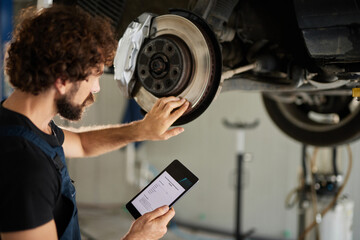 A mechanic inspects a brake disc while using a tablet in a car repair workshop. The worker is focused on the task, ensuring vehicle safety in early afternoon.