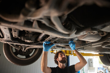 A skilled car mechanic conducts repairs on a vehicle from below in an auto repair shop. Tools are in use as the mechanic carefully inspects and works on the car's undercarriage.