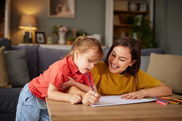 Mother and daughter share a joyful moment as they draw and color at a wooden table in their cozy living room filled with warmth and light.