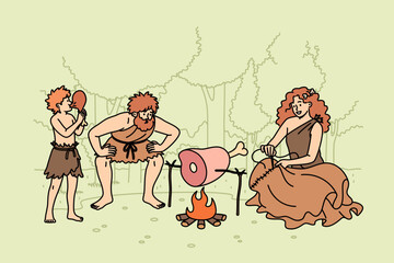Family of primitive people stands around fire in forest, roasting meat for dinner or sewing up clothes. Group of primitive people of different ages living in stone age dressed in rags