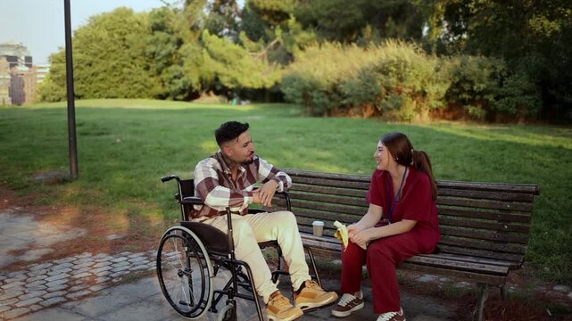 Friendly female nurse sitting on a park bench offers a banana to a young disabled man in a wheelchair. Compassionate caregiver providing support and assistance during a relaxing break outdoors