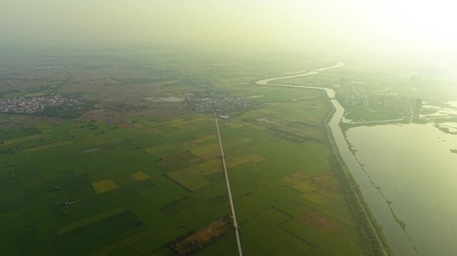 Aerial drone shot wide panning shot of fertile farmland landscape with lush fields, water body, and suburban surroundings at monsoon sunrise in Delhi, India.