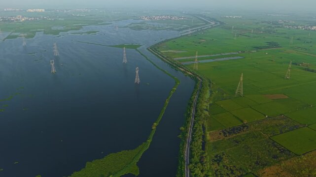 Aerial drone shot wide panning shot of lush green farmland and a calm water body with power cables stretching across, captured during sunrise in the monsoon season on the outskirts of Delhi, India.