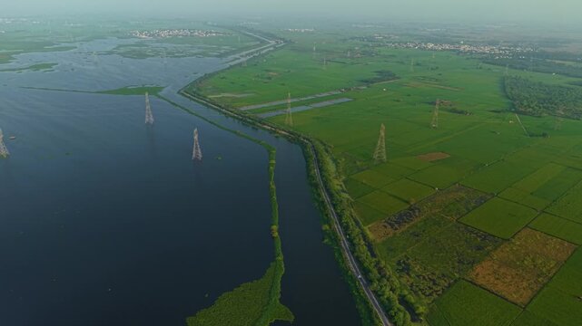 Aerial drone shot wide panning shot showcasing vibrant monsoon farmland and water body surrounded by power lines, glowing in sunrise light near Delhi, India.
