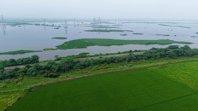 Aerial drone shot wide panning shot highlighting lush farmland and shimmering water body with power cables, monsoon season sunrise creating a soft rural mood near Delhi, India.