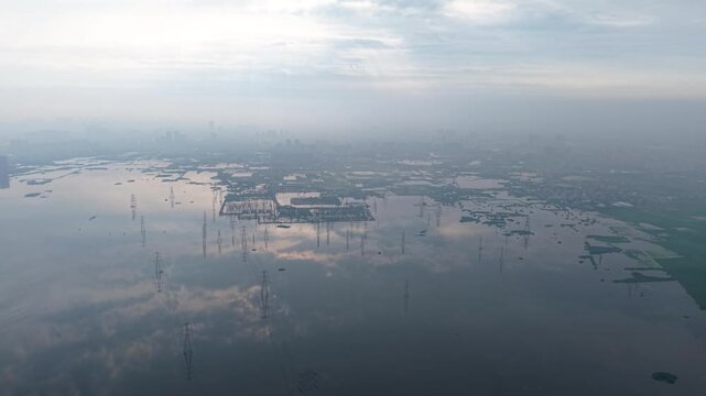 Panning aerial shot of water body and power lines stretching across, reflection visible on the calm surface, taken at monsoon sunrise near Delhi, India.