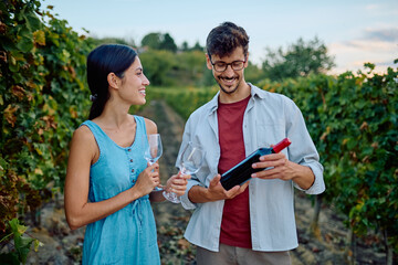 Happy young adult couple enjoying a date outdoors, holding wine and looking at the bottle while visiting a vineyard