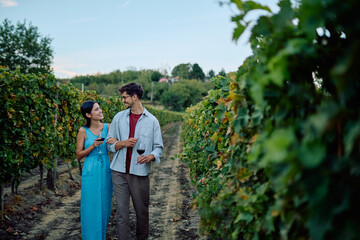 Smiling young couple enjoying an afternoon stroll through a lush vineyard at sunset, holding glasses of red wine
