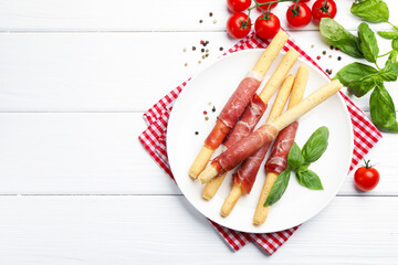 Delicious grissini sticks with prosciutto, peppercorns, tomatoes and basil on white wooden table, flat lay. Space for text