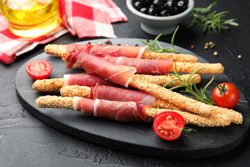 Delicious grissini sticks with sesame seeds, prosciutto, rosemary and tomatoes on dark textured table, closeup