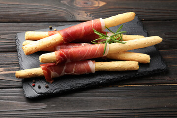 Delicious grissini sticks with prosciutto, peppercorns and rosemary on wooden table, closeup
