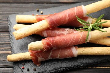 Delicious grissini sticks with prosciutto, peppercorns and rosemary on wooden table, closeup