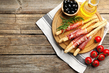 Delicious grissini sticks with sesame seeds, prosciutto and other products on wooden table, flat lay. Space for text