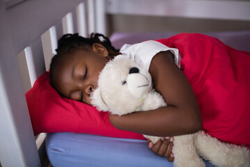 African American child sleeping in bedroom on bed frame hugging white teddy bear under red blanket