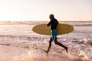 Senior African American man in swim shorts carrying yellow surfboard wading on beach at sunset