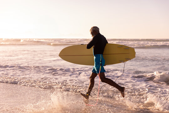 Senior African American man in swim shorts carrying yellow surfboard wading on beach at sunset