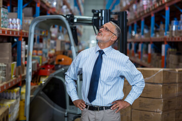Senior man wearing shirt and tie inspecting shelves with forklift, wrapped pallets in warehouse
