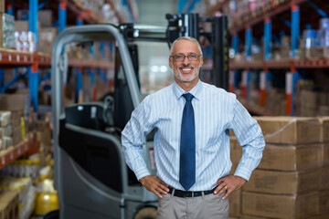 Senior manager standing with hands on hips in warehouse, with forklift, racks and cardboard boxes