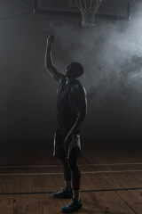 Lone athlete standing under basketball hoop on misty wooden court floor, casting subtle shadows