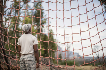 African American man in camouflage standing facing orange rope cargo net in forest training ground