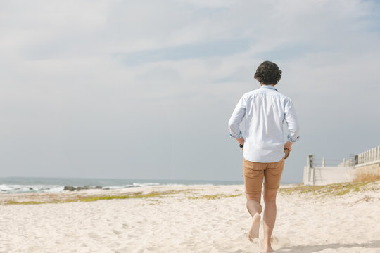 Man walking barefoot toward ocean waves on beach by boardwalk ramp with wristwatch, copy space
