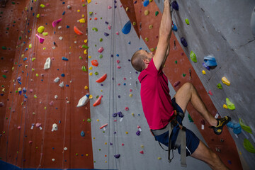 Adult male climber scaling wall at gym using harness rope and specialized shoes on colorful holds