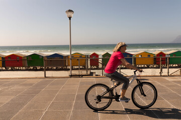 Senior woman riding bicycle along coastal path passing railing, colorful huts and lamp, copy space