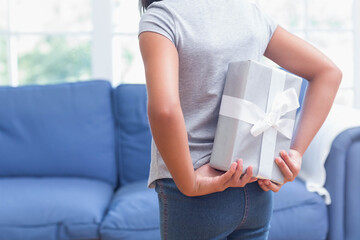 Mid adult woman holding silver gift box behind her back at home with sofa, copy space
