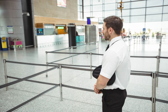 Male pilot wearing uniform standing near queue barriers at airport terminal holding cap, copy space - Powered by Adobe